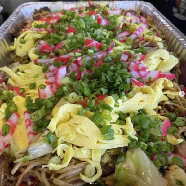 A large foil pan of Hawaiian noodles topped with green onions, egg strips, and pink and white fish cake.