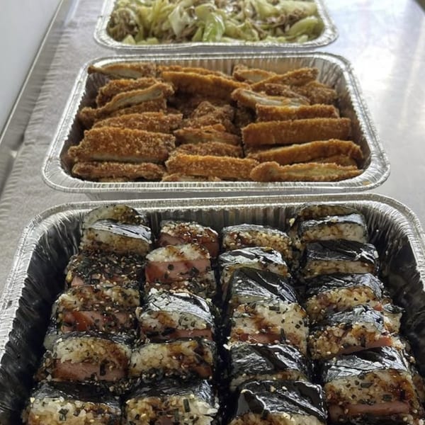 Three foil trays of Hawaiian food, including spam musubi, fried chicken, and cabbage.